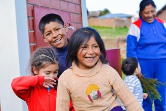 niños de bajos recursos posando felices para la cámara. Atrás, una señora sonríe a un niño mientras sostiene en sus manos algunas plantas.