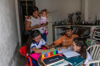 Dos niñas y su papá hacen tareas felices mientras su mamá los mira y carga un bebé en sus brazos 
